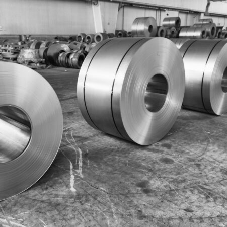 Large stainless steel coils made from rolled steel strip stored in an industrial hall, raw material for the manufacture of process vessels and industrial plant components.